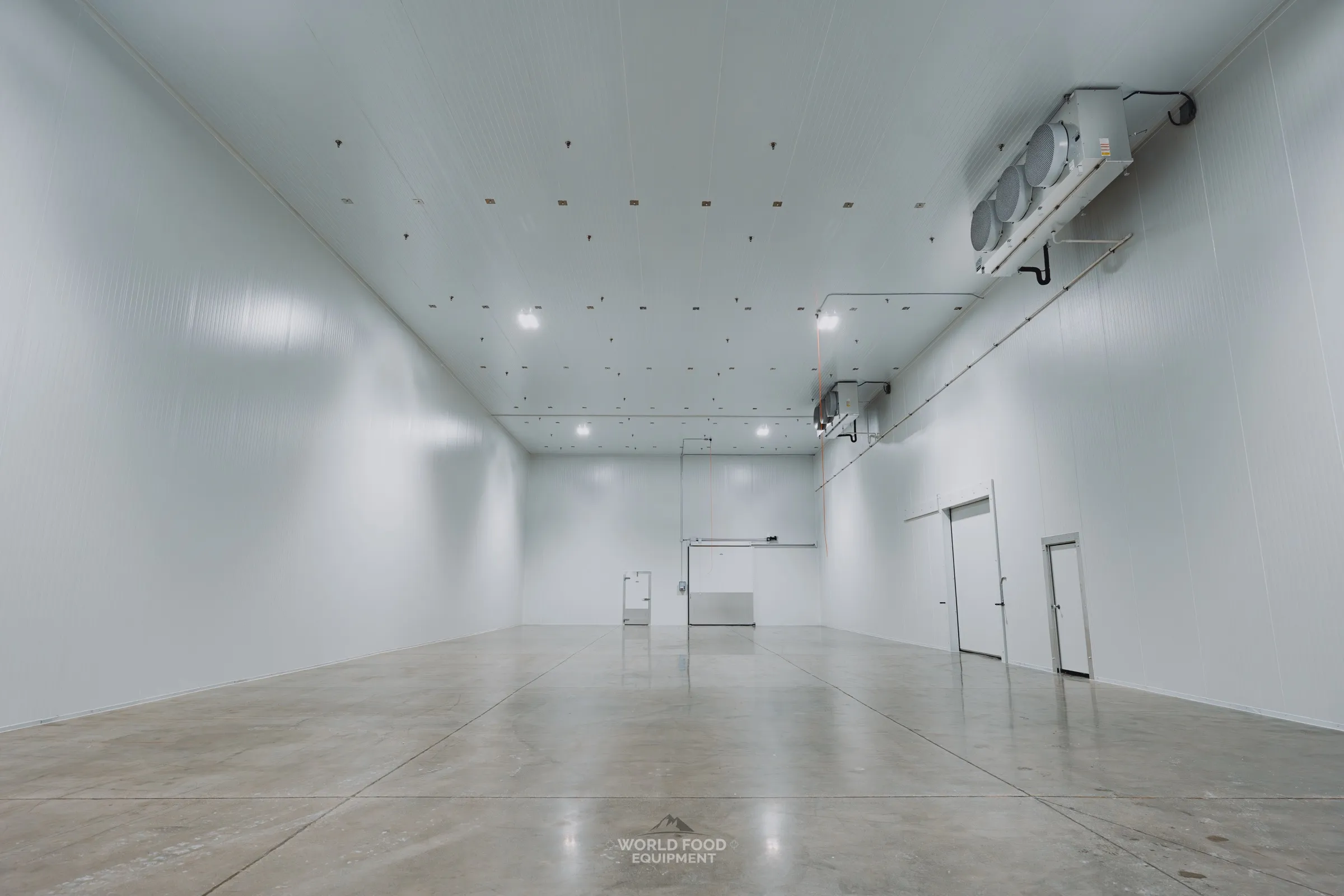Wide insulated cold storage room with white panel walls, polished concrete floor, ceiling-mounted refrigeration evaporators, and sliding insulated doors at the end, Ontario commercial installation by World Food Equipment