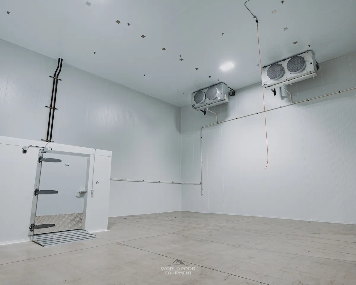 Finished commercial cold storage room with white insulated panel walls, an insulated swing door with diamond-plate kickplate on the left, a two-fan ceiling-mounted evaporator on the right wall, polished concrete floor, Ontario commercial cold storage by World Food Equipment