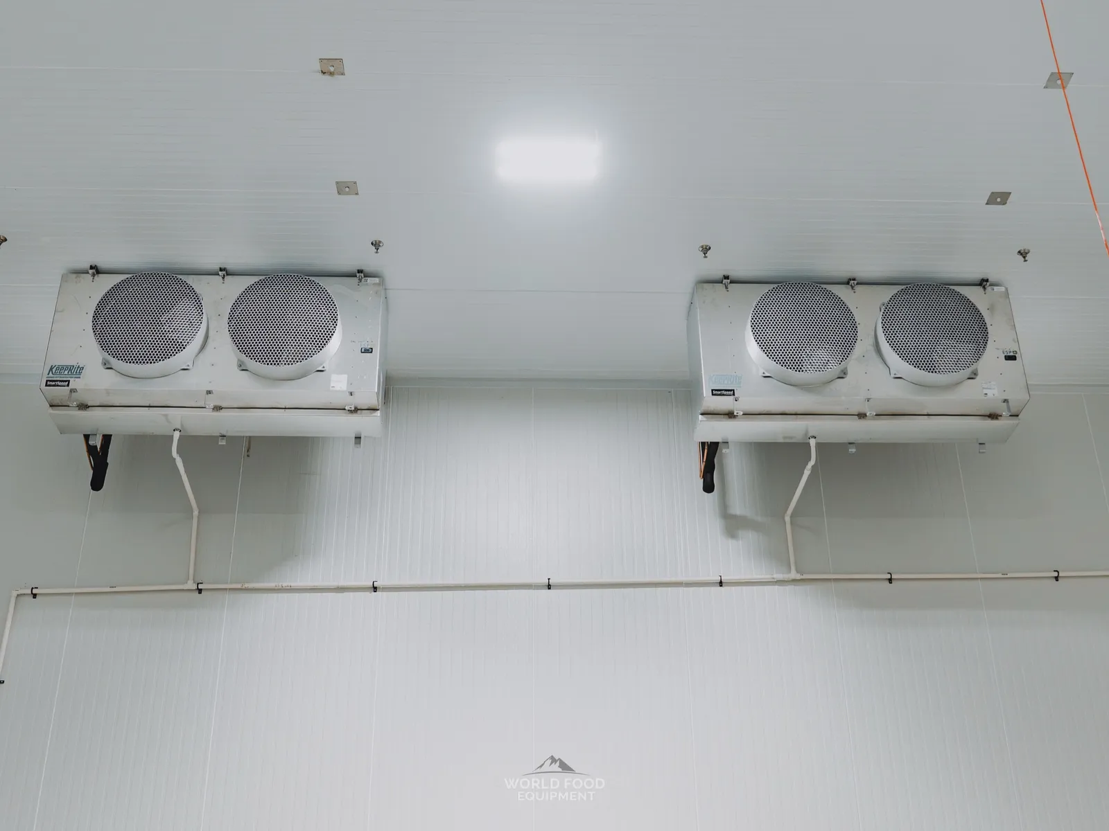 Two KeepRite SmartSpeed two-fan ceiling-mounted evaporators side by side on the same wall at the Pacer Air Freight medical-grade cold storage in Mississauga, refrigeration lines routed between the units along the white insulated panel wall above, pharma-grade multi-zone refrigeration sized to the combined cooler-plus-freezer load.