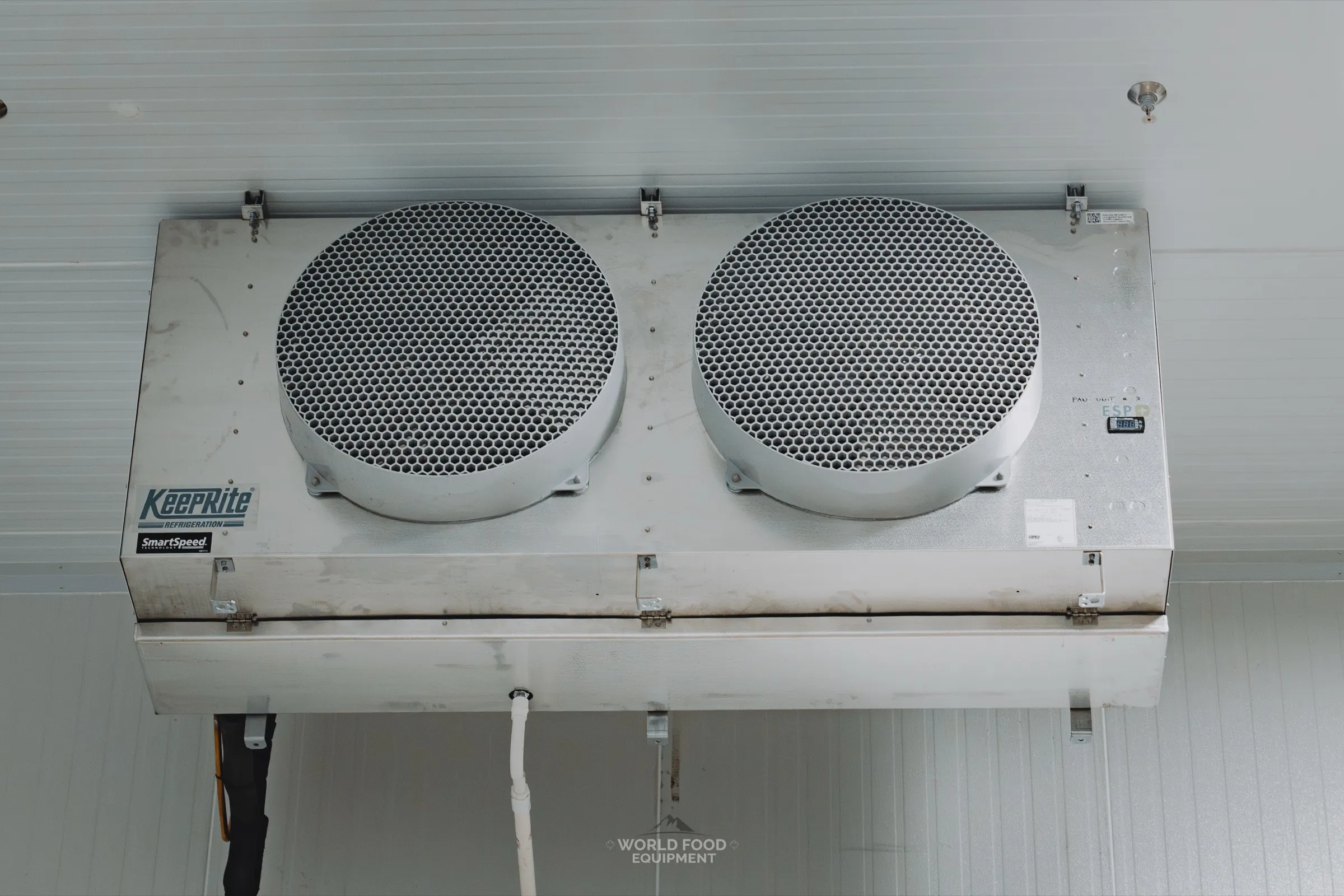 Close-up of a single KeepRite SmartSpeed evaporator unit ceiling-mounted in a finished operational walk-in, brand decal visible on the front housing, dual fan grilles, refrigerant line set with foam insulation routed below, ceiling sprinkler head, Pacer Air Freight install, Ontario, World Food Equipment.