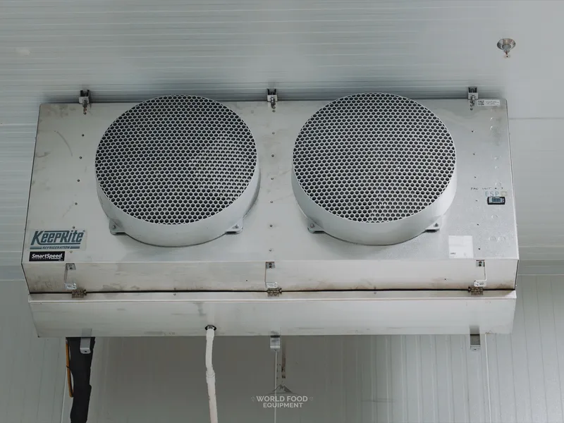 Close-up of a KeepRite Refrigeration SmartSpeed two-fan ceiling-mounted evaporator at the Pacer Air Freight medical-grade cold storage in Mississauga, KeepRite Refrigeration logo and SmartSpeed branding visible on the unit casing, refrigeration drain line wrapped in black insulation tape running below.
