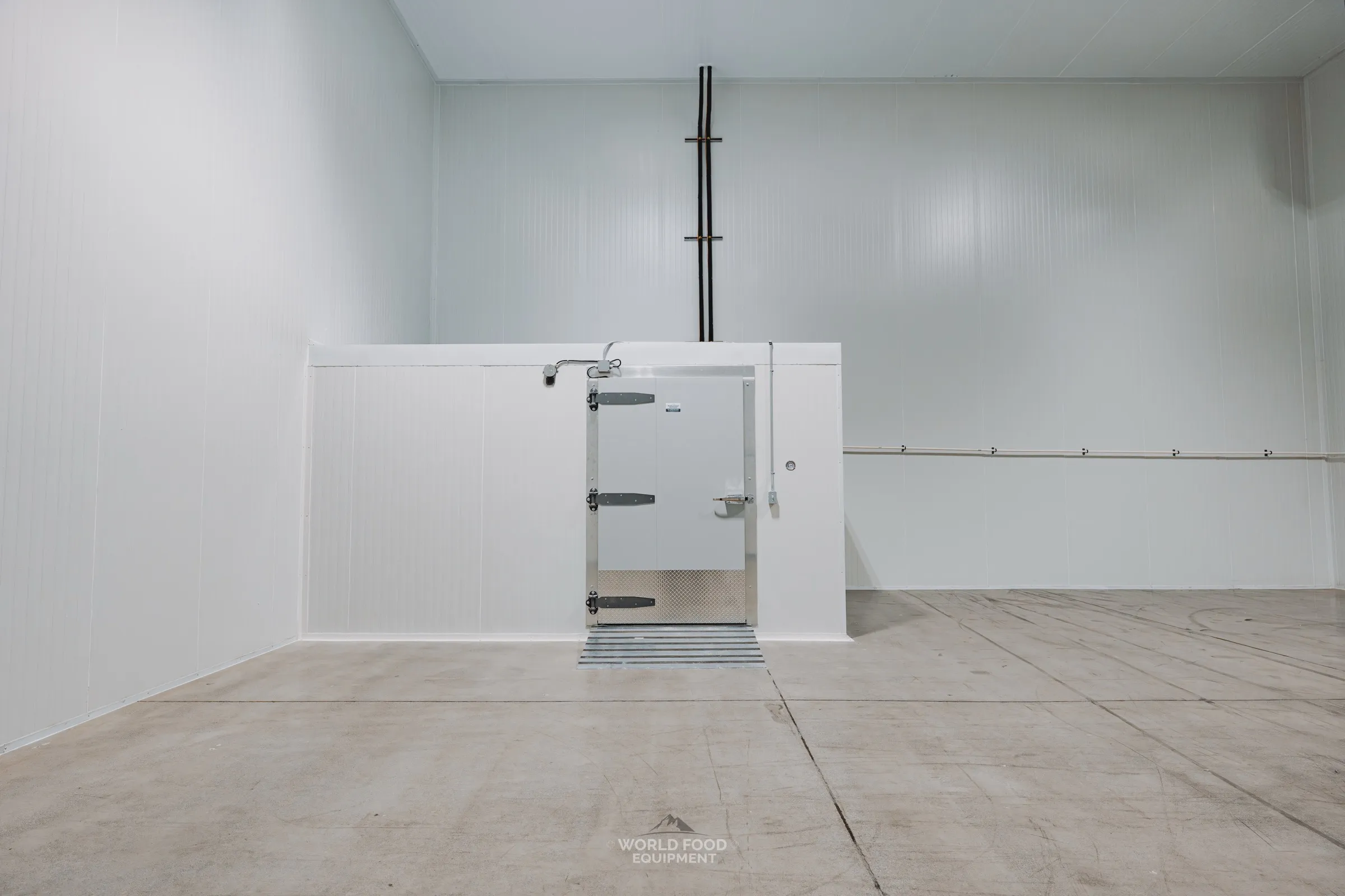 Compact standalone commercial walk-in cooler with white insulated panels, swing-style insulated door, and diamond plate threshold, sitting on a finished concrete floor inside an open warehouse, Pacer Air Freight install, Ontario, World Food Equipment