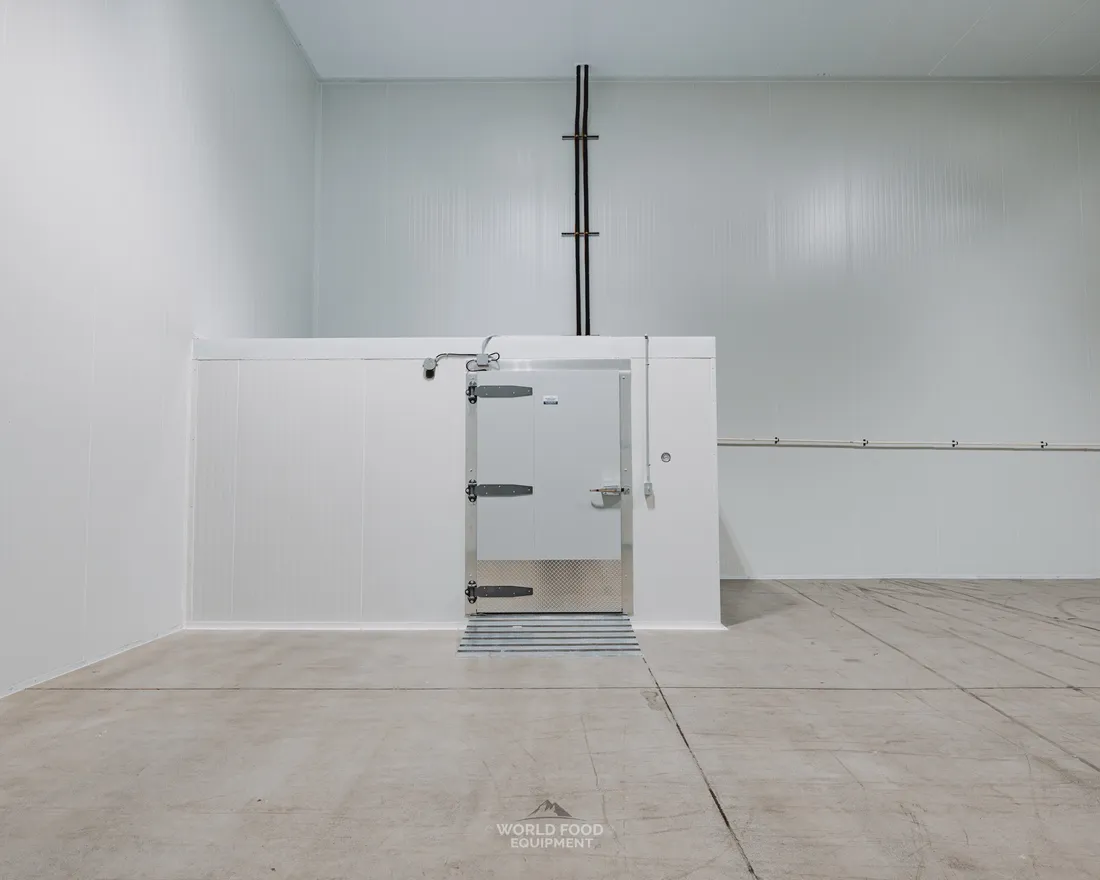 Insulated cold-room swing door with multi-point latch hardware and heated threshold strip on the floor, set into a white insulated panel wall in a mid-size cold storage room, Pacer Air Freight install, Mississauga, World Food Equipment