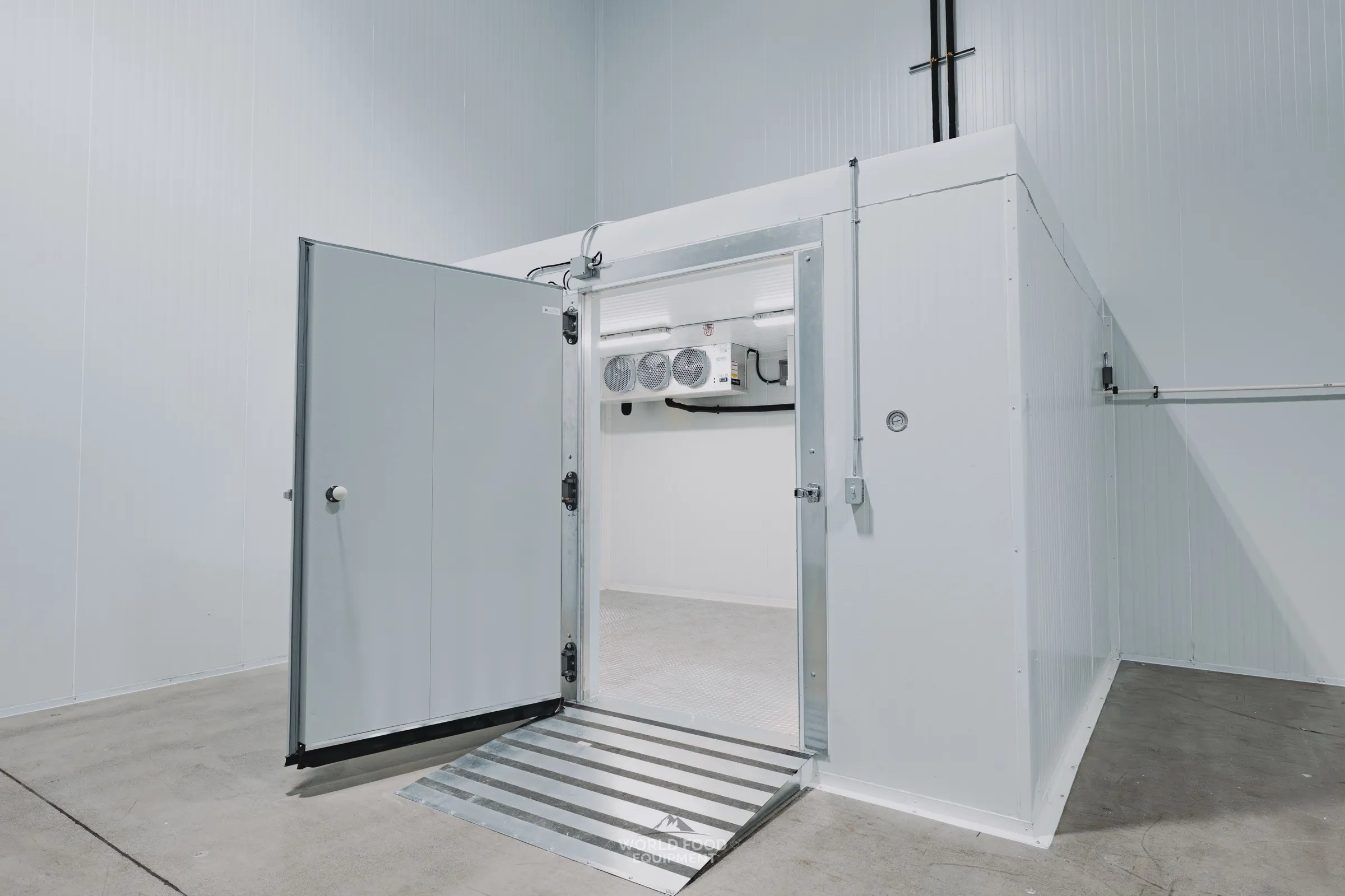 Finished commercial walk-in cooler with the insulated swing door open showing a diamond-plate ramp leading into a smaller cold storage room, ceiling-mounted evaporator visible inside, white insulated panel walls, Pacer Air Freight install at the WFE Mississauga HQ service area, Ontario commercial walk-in installation by World Food Equipment