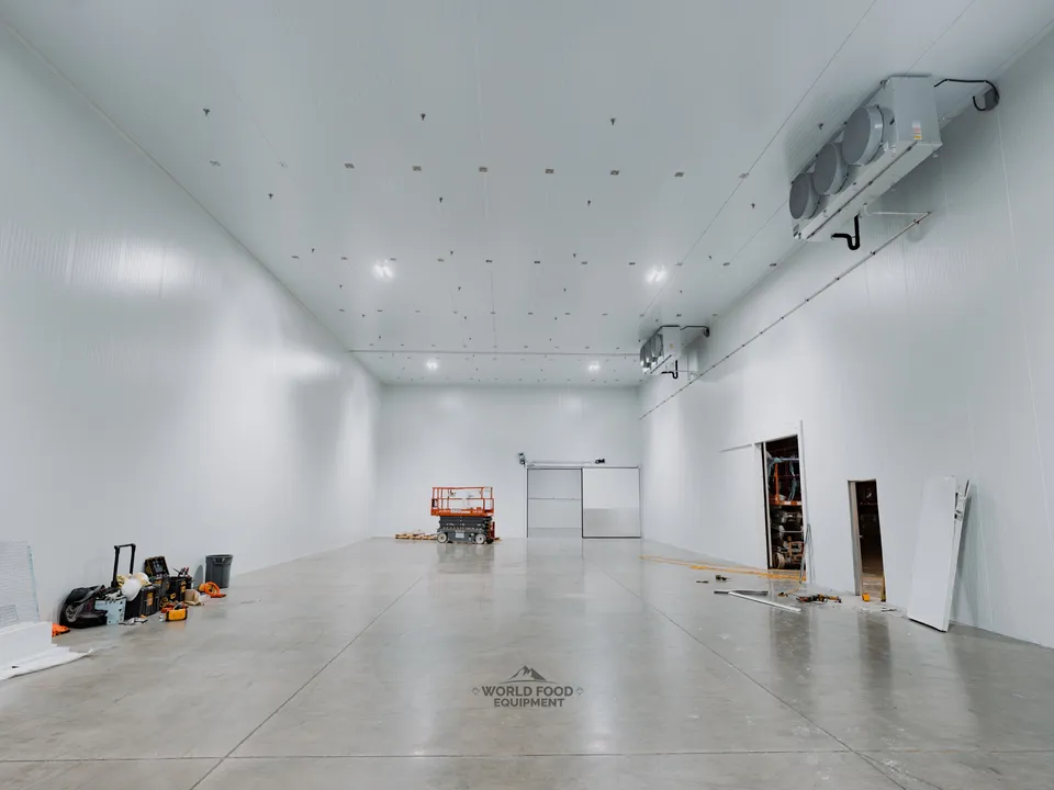 Cathedral-scale interior of the Pacer Air Freight medical-grade cold storage envelope in Mississauga with refrigeration installed: KeepRite ceiling-mounted evaporator hung against the right wall, scissor lift staged in the distance, finishing-phase debris near the doorways at the far end.