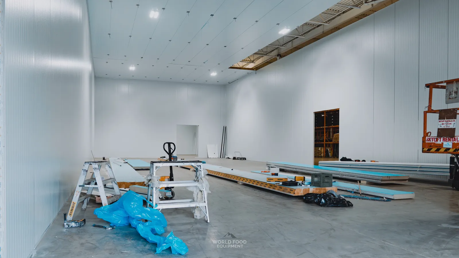 Cathedral-scale industrial cold storage envelope mid-build with insulated panels staged on the floor, sawhorses and step ladder set up for panel cutting, blue protective tarps over staged materials, white panel walls and ceiling already installed but refrigeration not yet tied in, Pacer Air Freight install, Ontario, World Food Equipment