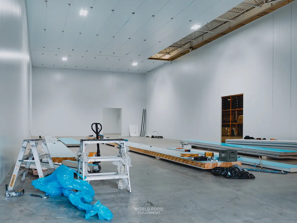 Mid-build interior of a smaller compartment at the Pacer Air Freight medical-grade cold storage envelope in Mississauga: scaffold and ladder staged mid-room, insulated panel sections lying on the polished concrete floor mid-construction, white insulated panel walls and ceiling already in place around the room.