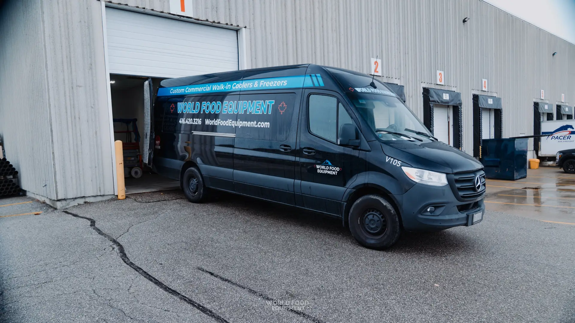 World Food Equipment-branded black Mercedes Sprinter van at a commercial loading bay, WFE wordmark and worldfoodequipment.com across the rear quarter-panel, parked at the Pacer Air Freight cold storage install site in Mississauga, Ontario.