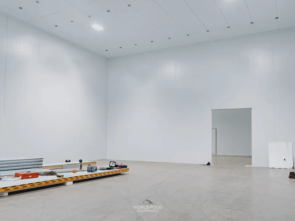 Cathedral-scale interior of the Pacer Air Freight medical-grade cold storage envelope in Mississauga with envelope walls and ceiling nearly complete, Norbec-branded insulated panels staged on the polished concrete floor for the next pharma-grade zone, doorway through to an adjacent smaller room visible at the far end.