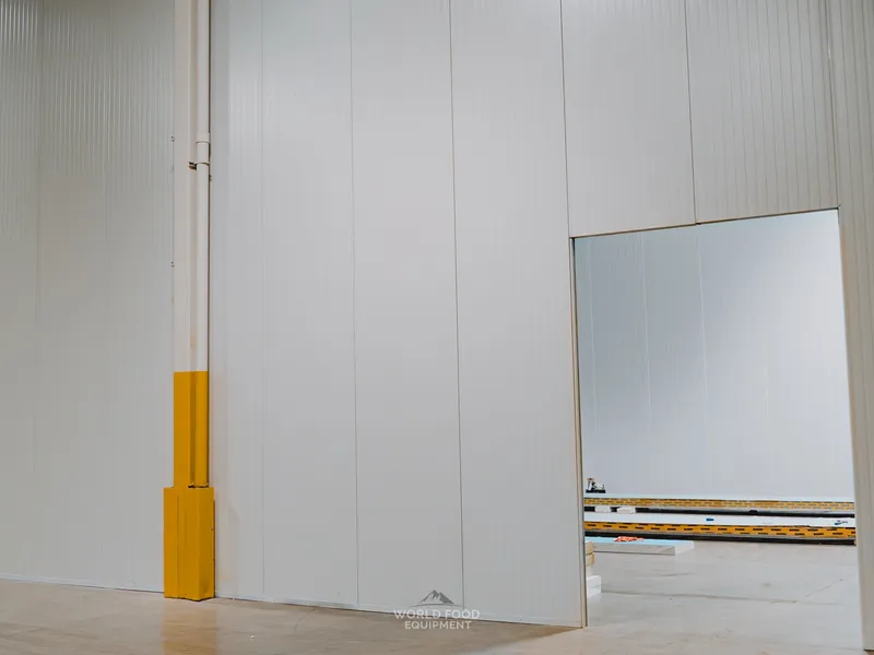 Exterior view of the Pacer Air Freight medical-grade cold storage envelope being built in Mississauga with yellow protective sleeving wrapped around a building structural support column where the envelope walls meet the existing structure, doorway through to an adjacent smaller cold room visible.