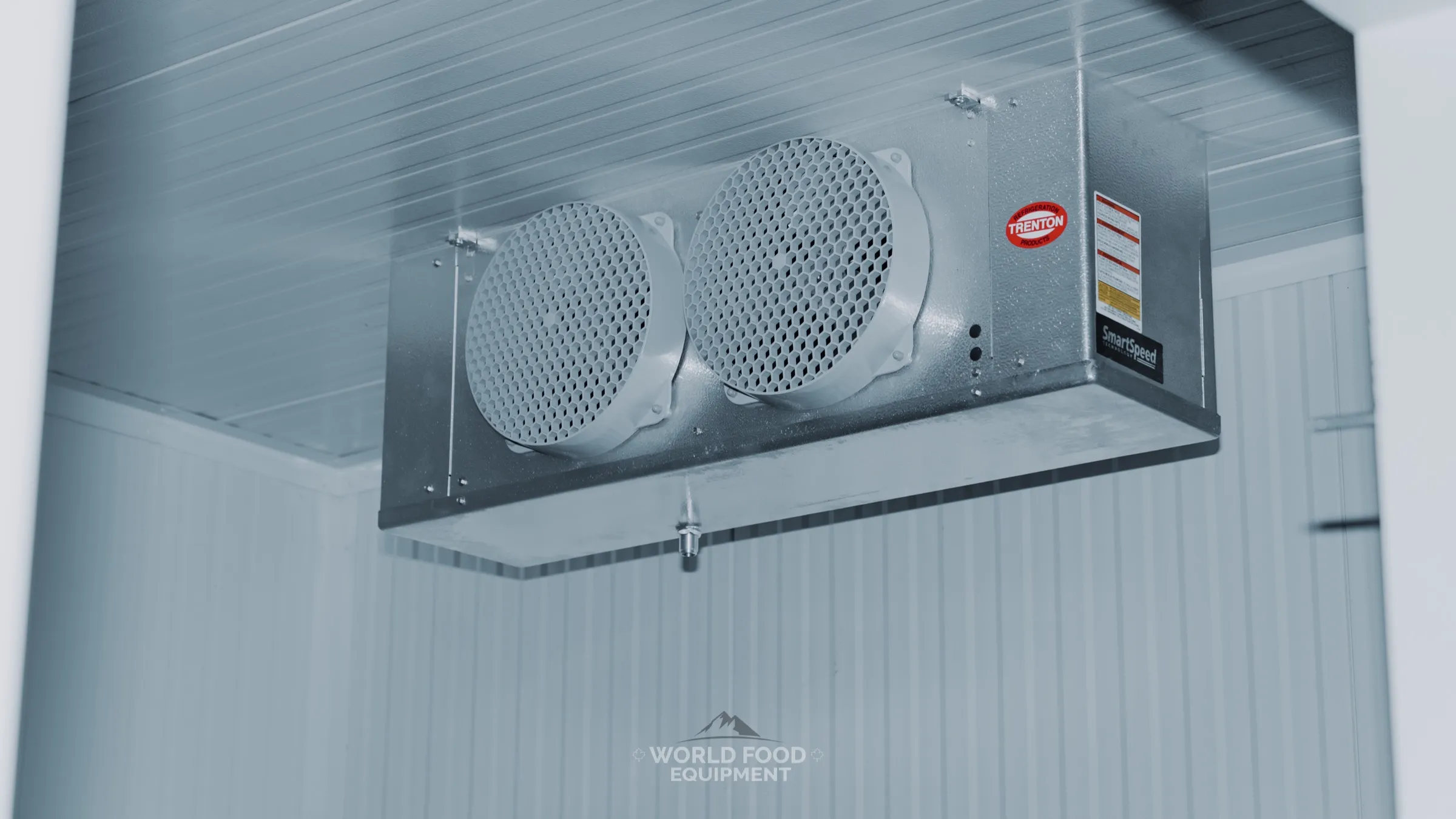 Trenton SmartSpeed twin-fan evaporator ceiling-mounted in a finished white-panel walk-in at the Progress Avenue install in Ontario, condensate drain fitting on the underside of the pan, World Food Equipment