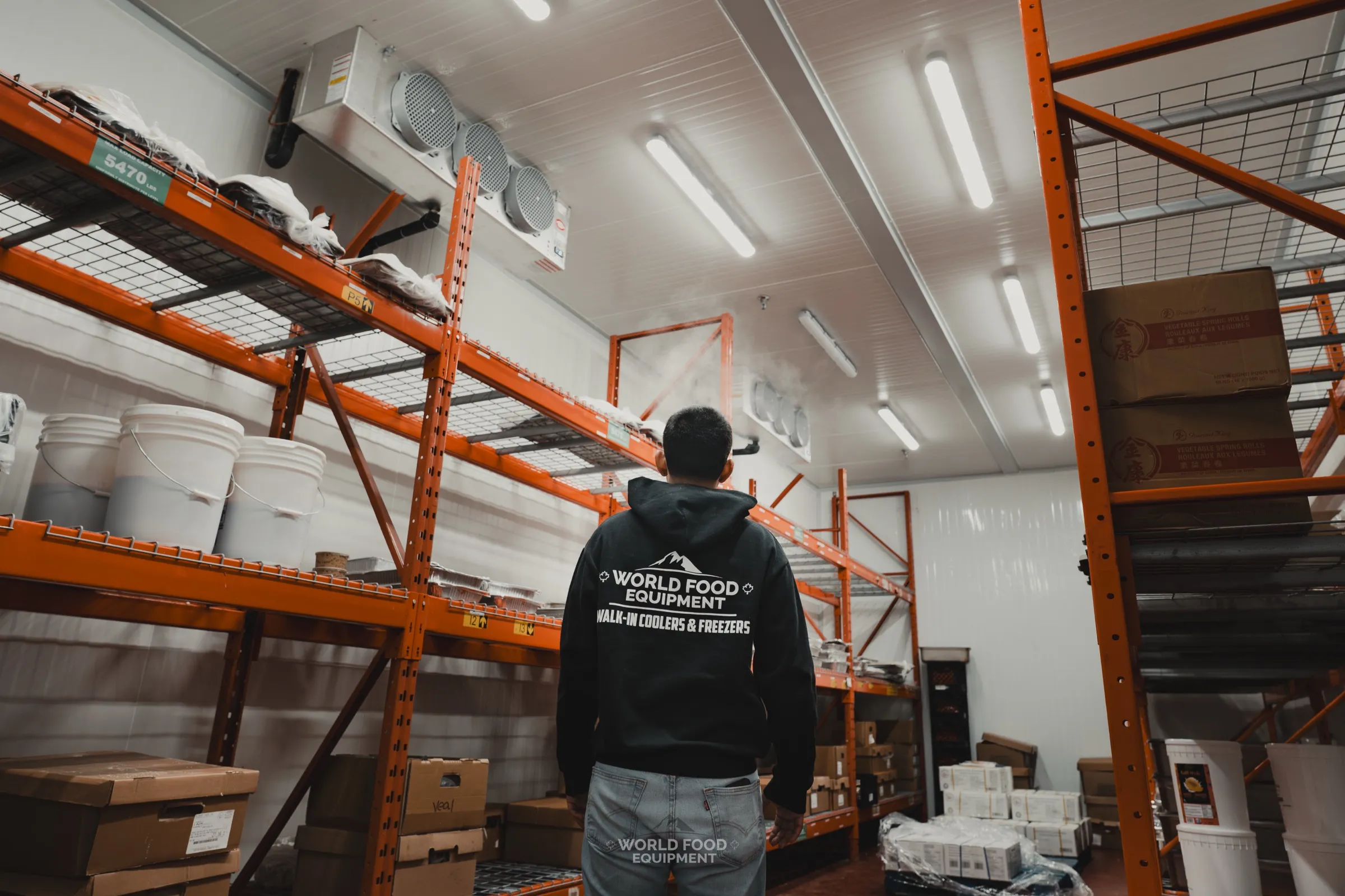Interior of the finished walk-in freezer at the Stainless Steel processing equipment fabricator. Orange industrial pallet racking loaded with inventory, ceiling-mounted commercial evaporator unit overhead, insulated panel walls and ceiling, World Food Equipment-branded crew member walking the aisle for scale