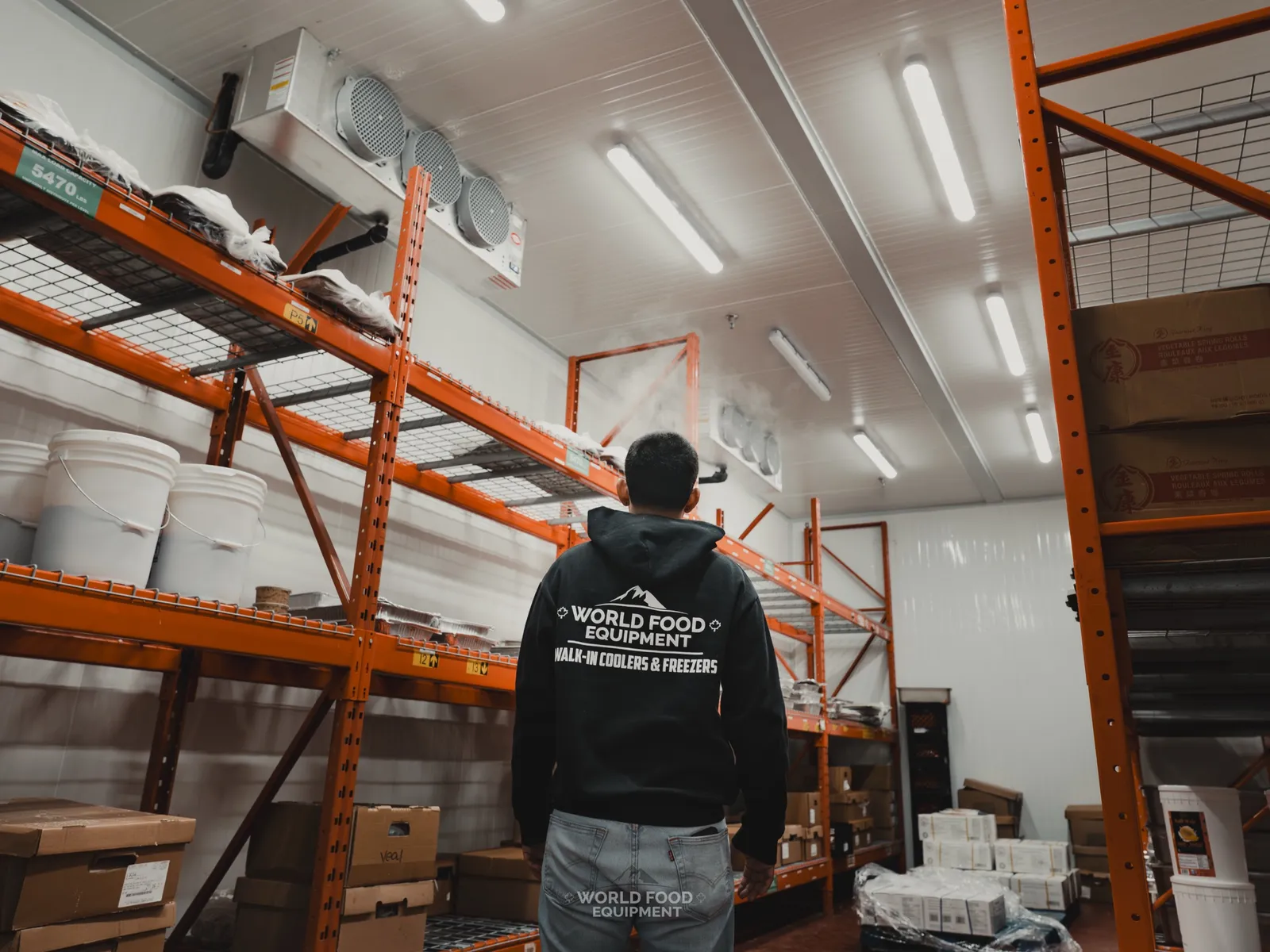 WFE installer in branded black hoodie inside one of the two finished Stainless Steel cold rooms with stocked product across the custom orange shelving on both sides and the ceiling-mounted evaporator visible overhead.