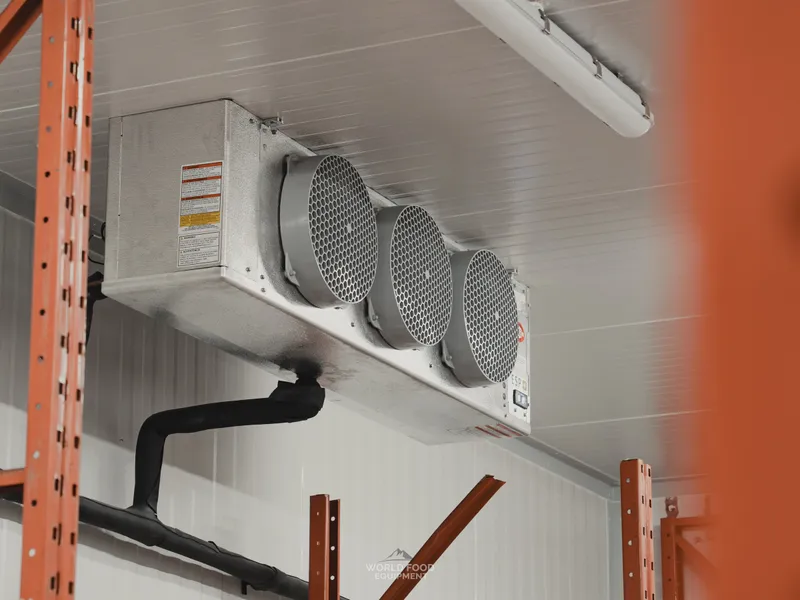 Three-fan ceiling-mounted evaporator inside a finished commercial cold storage room, orange pallet racking framing the view, insulated refrigeration drain piping running down the wall, white insulated panel walls, Stainless Steel install, Ontario, World Food Equipment
