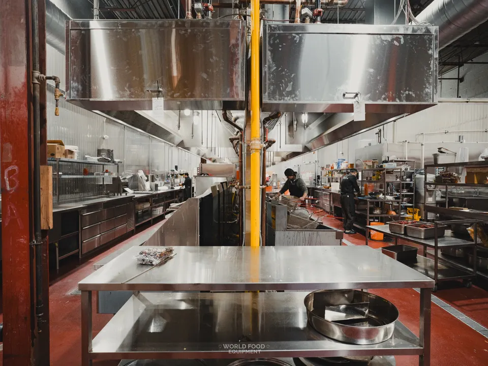 Finished commercial kitchen at Stainless Steel with fabricated stainless prep tables, a stainless cook line, twin exhaust hoods overhead, and kitchen staff working, red epoxy floor throughout.