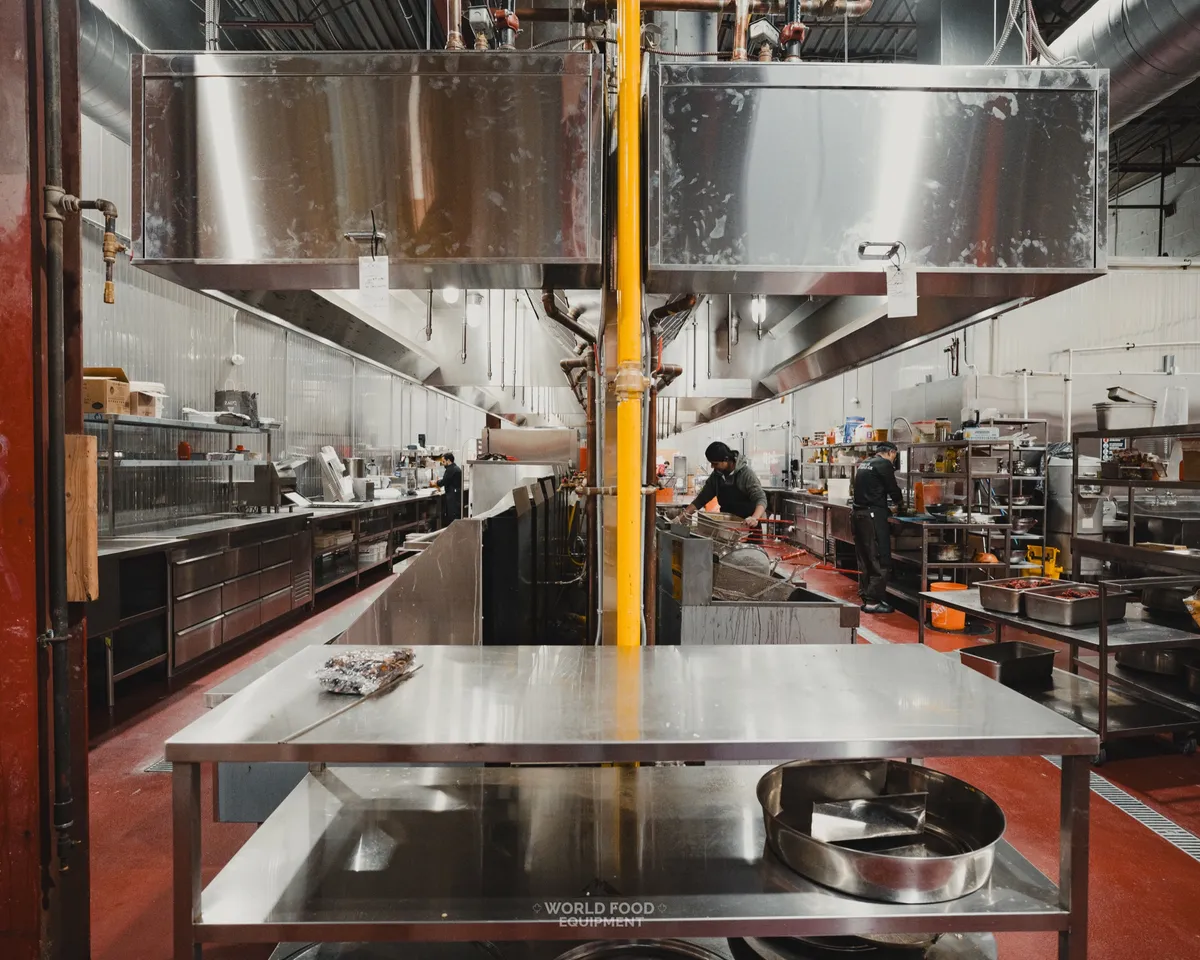 Busy commercial restaurant kitchen with stainless prep tables, two suspended stainless exhaust hoods, two cooks working on the line, red epoxy floor, Stainless Steel install, Ontario commercial kitchen by World Food Equipment
