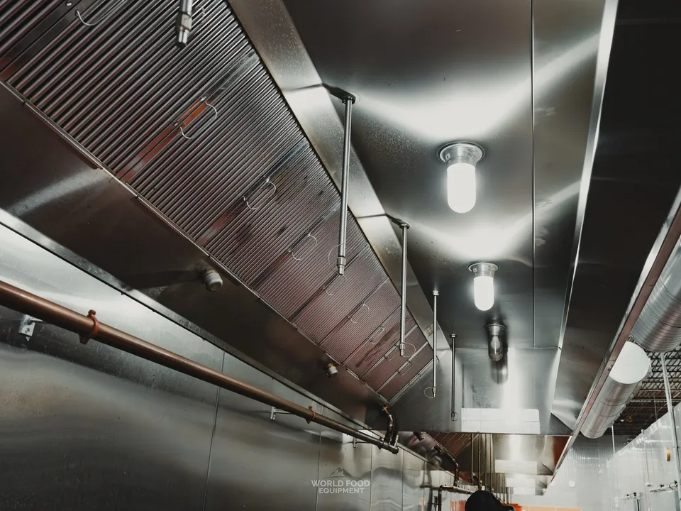 Commercial exhaust hood and ventilation canopy from below at the Stainless Steel kitchen, polished stainless with baffle filters, vapor-proof ceiling lights, and copper service lines run along the wall.