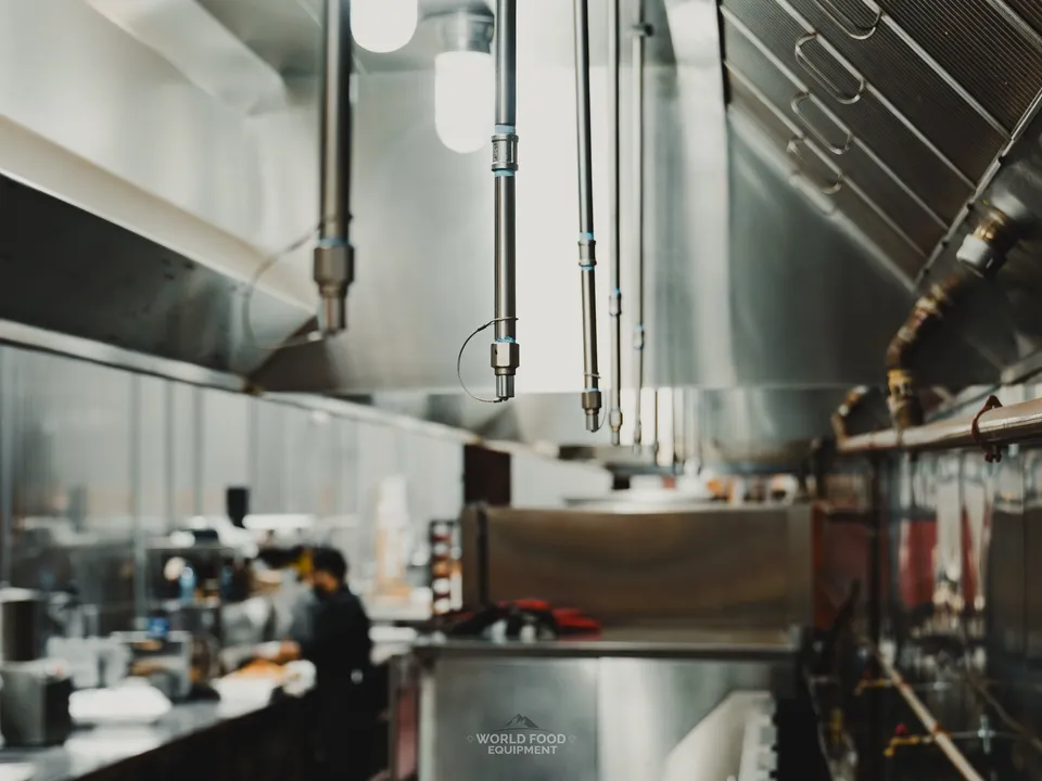 Close-up of the stainless commercial exhaust hood over the Stainless Steel cook line, baffle filters and ventilation built into the canopy, kitchen line visible behind.