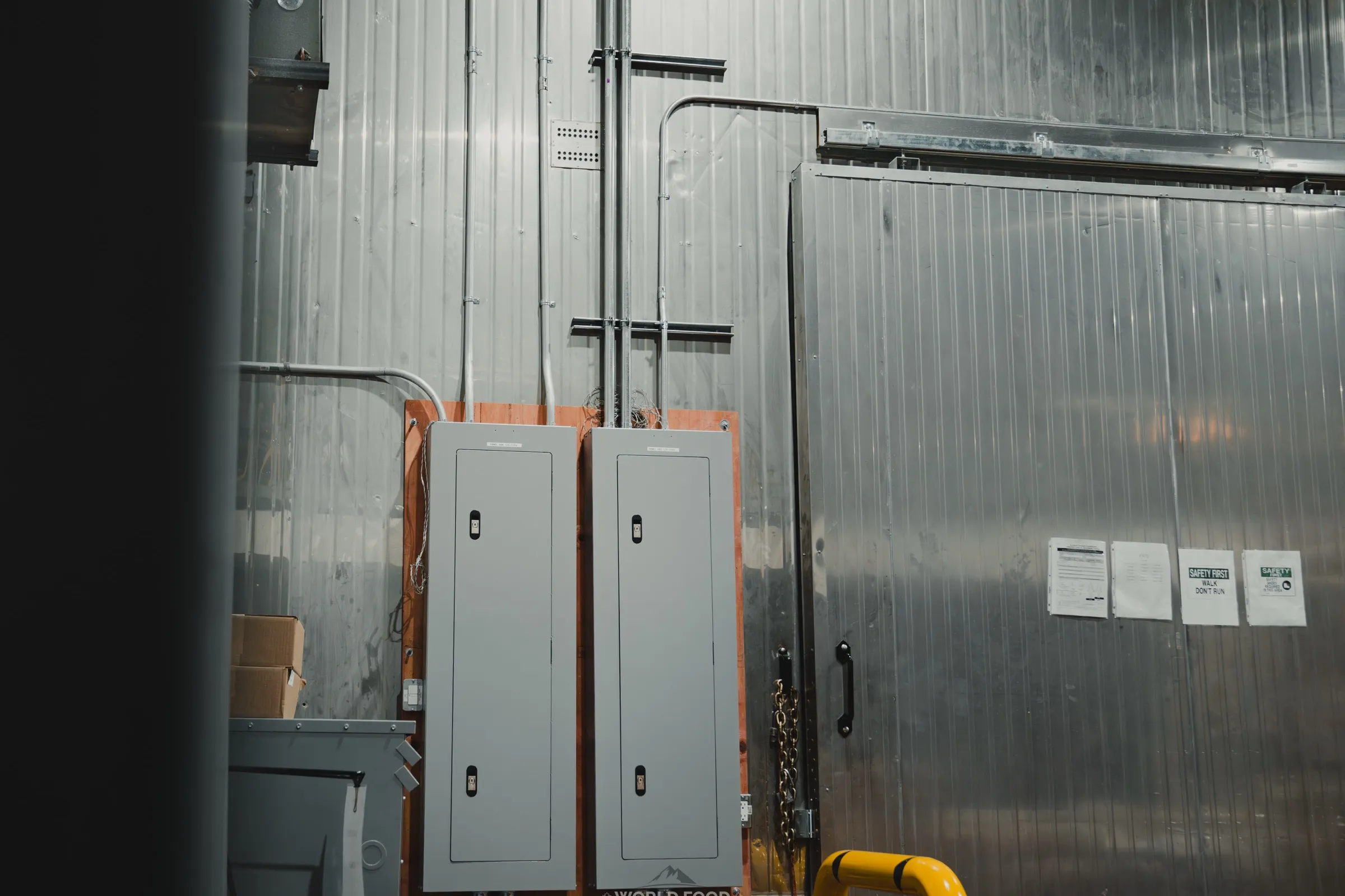 Two grey electrical distribution panels mounted on a plywood backboard beside a stainless-clad walk-in freezer door, EMT conduit runs rising to the ceiling, at the Stainless Steel project in Ontario, World Food Equipment