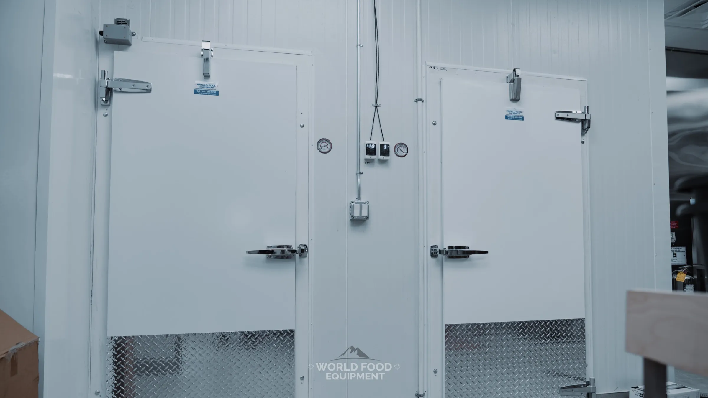 Close-up of twin walk-in cooler doors at The Bagel House restaurant install: white insulated doors with magnetic gaskets, stainless cam-locking handles, diamond-plate kickplates at the base, analog temperature gauge centered between them, World Food Equipment Ontario restaurant refrigeration