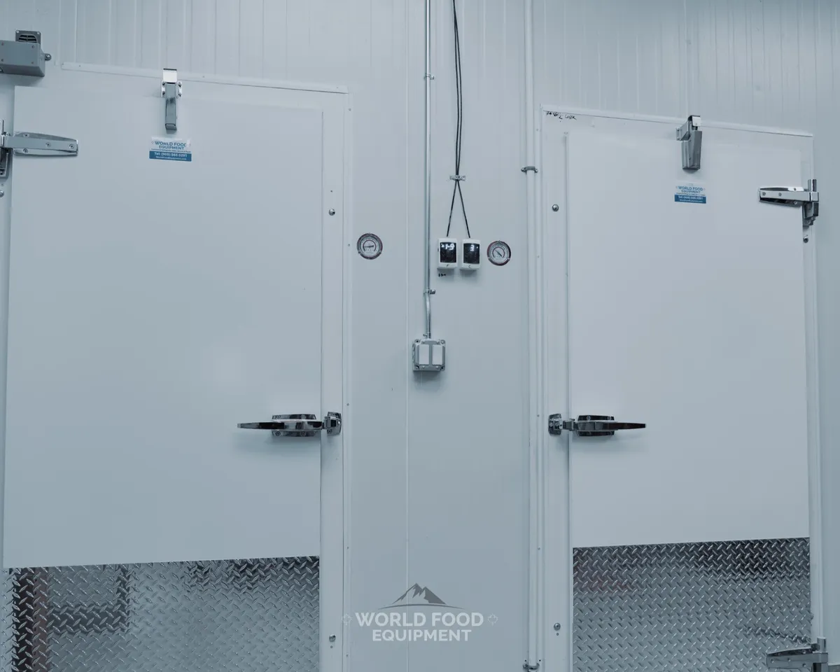Two finished white insulated walk-in cooler and freezer doors side by side with diamond-plate kickplates, dual latch handles, NSF dial thermometer and digital temperature controllers mounted between them, bright commercial bakery back-of-house, The Bagel House install, Ontario commercial walk-in installation by World Food Equipment