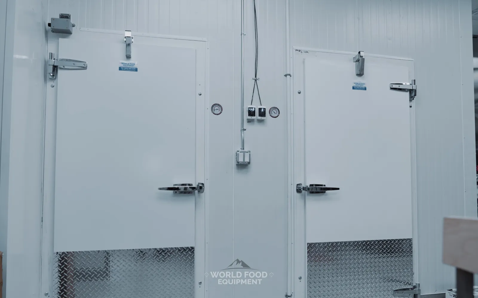 Twin commercial walk-in cooler swing doors with white insulated panels, heavy-duty cam-lock latches, diamond-plate kickplates, and two wall-mounted digital temperature controllers with an analog backup gauge between the doors, The Bagel House install, Ontario, World Food Equipment