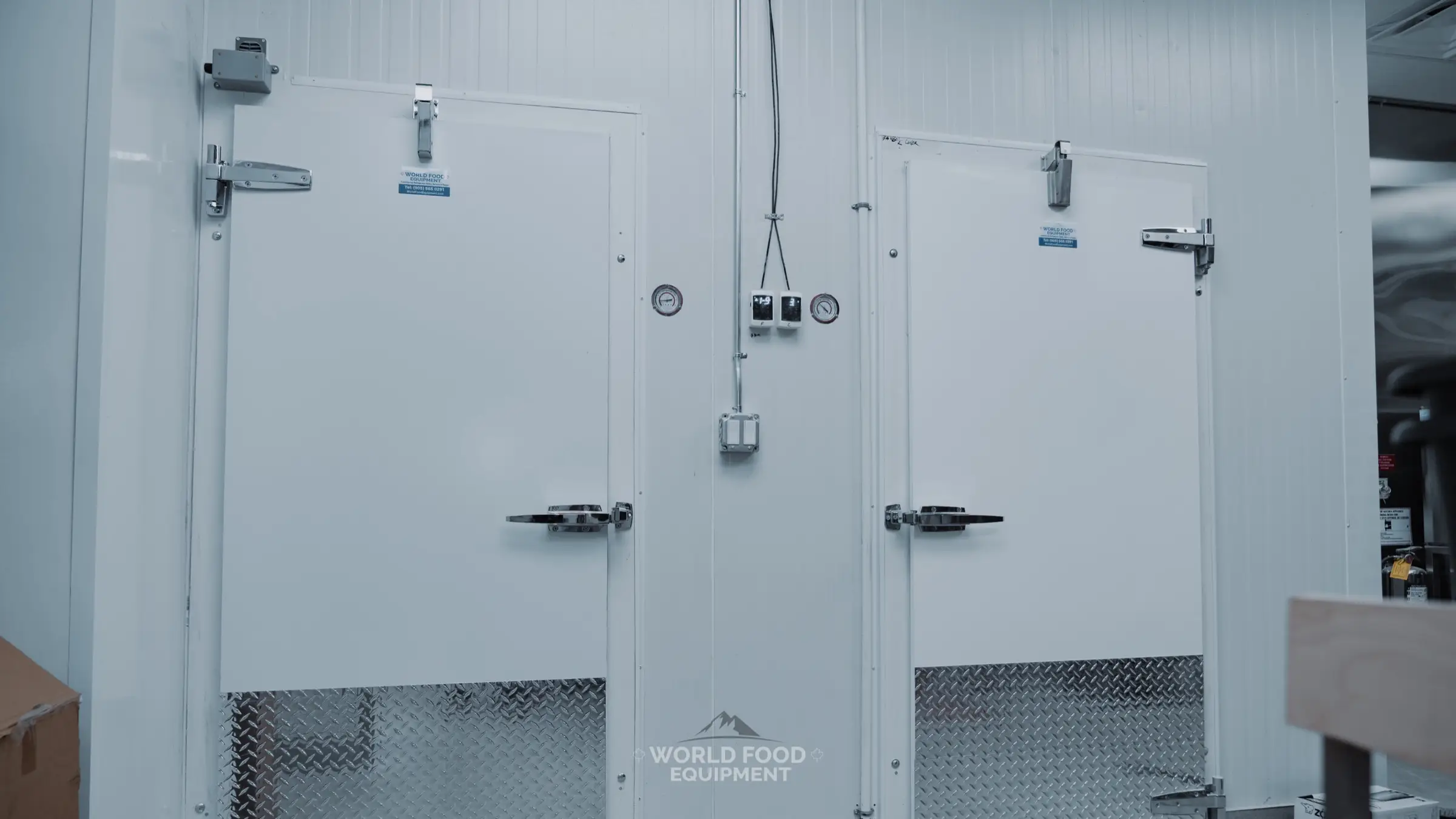Twin white walk-in cooler and freezer swing doors at The Bagel House in Toronto, chrome hinges and latches, diamond-plate kickplates, two Dixell digital controllers and dial thermometers on the panel between the doors, WFE install.