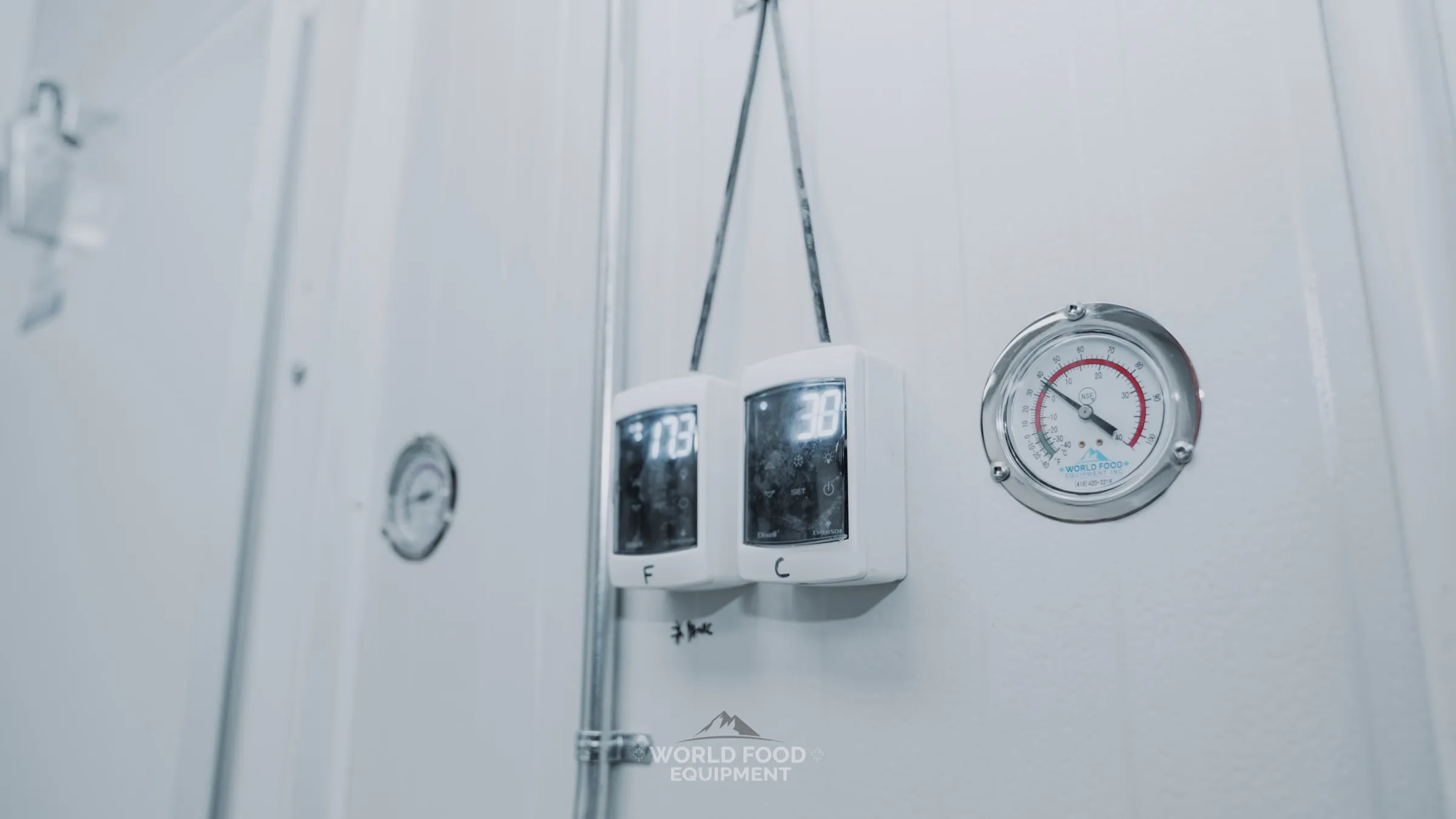 Close-up of a WFE-branded NSF stainless dial thermometer mounted on a white insulated walk-in panel wall, with two Dixell digital temperature controllers hanging beside it on a coiled cable showing live Fahrenheit and Celsius readings, The Bagel House install, Ontario, World Food Equipment