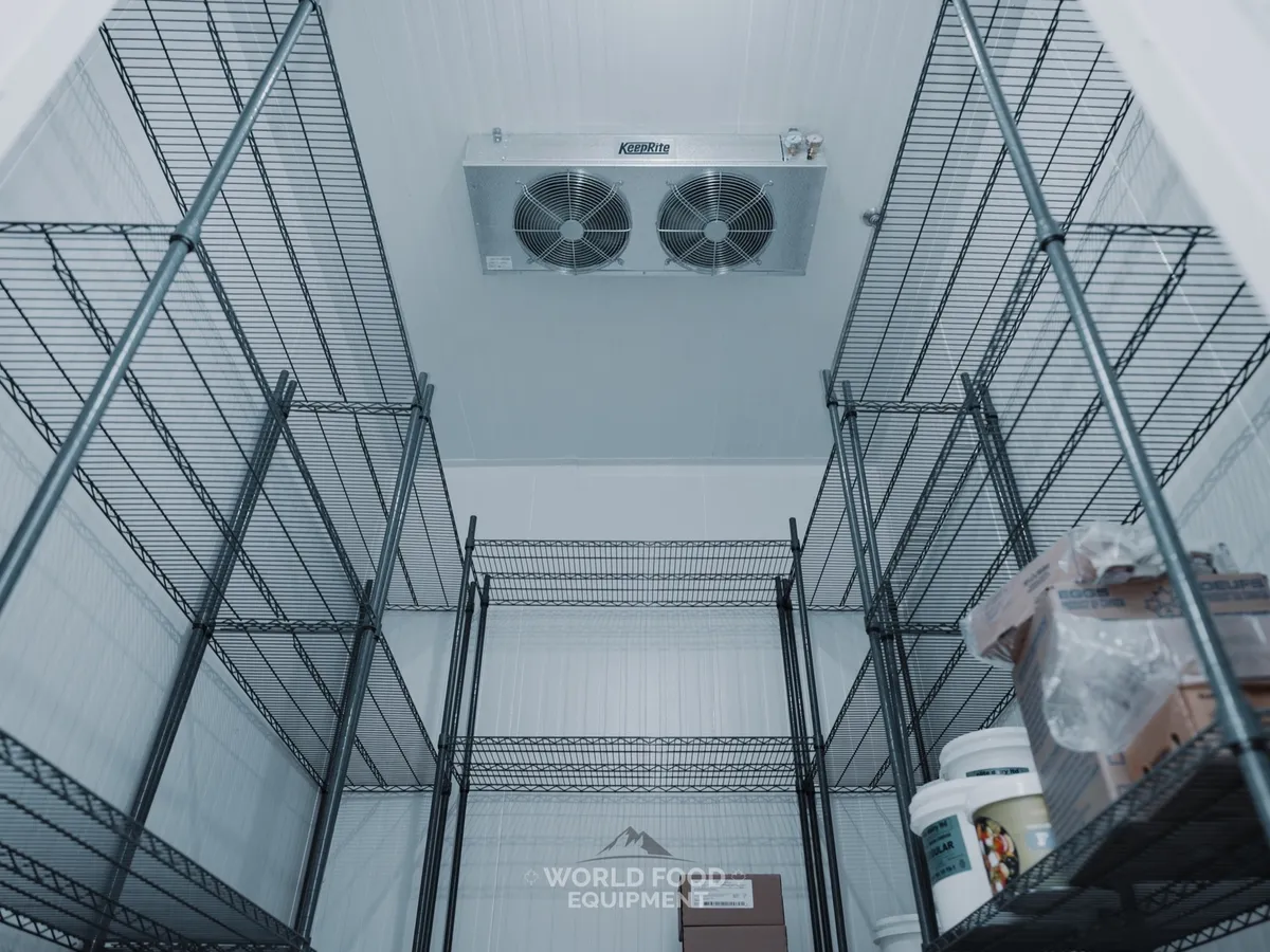 Inside The Bagel House walk-in looking up at the KeepRite twin-fan ceiling evaporator, black wire shelving down both sides, egg cases and dairy tubs stocked on the lower shelves, white insulated panel walls and ceiling.