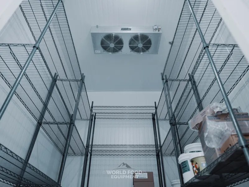 Commercial walk-in cooler interior viewed from a low angle, black wire storage shelving lining both walls, a ceiling-mounted twin-fan evaporator overhead, white insulated panel walls, stocked product on the shelves, The Bagel House install, Ontario, World Food Equipment