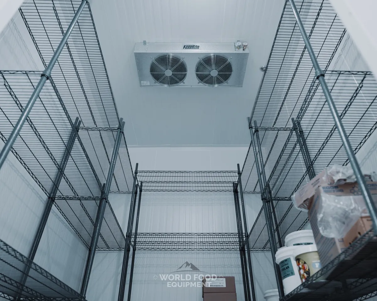 Interior of a finished commercial walk-in cooler looking up toward a ceiling-mounted twin-fan evaporator, wire shelving loaded with product on both sides, white insulated panel walls, The Bagel House install, Ontario commercial walk-in cooler by World Food Equipment