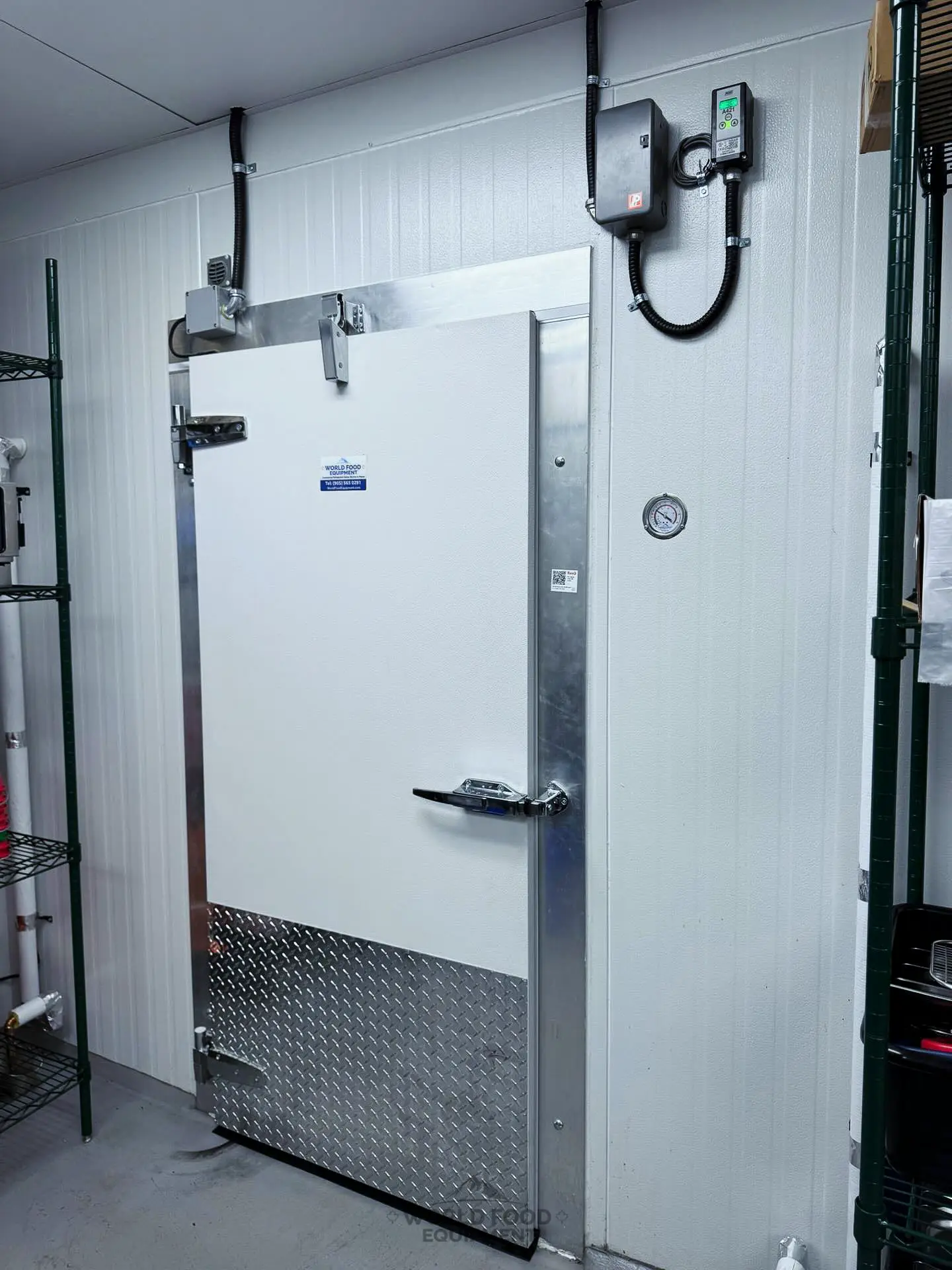 Real World Food Equipment walk-in cold-room swing door: aluminium-wrapped insulated leaf with cam-lift self-closing hinges, magnetic perimeter gasket, safety-release latch handle, diamond-plate kickplate, and a wall-mounted digital temperature controller with analog dial gauge