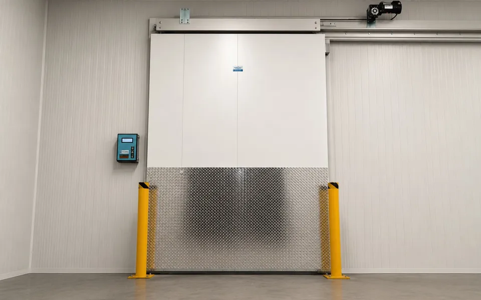 Finished commercial walk-in cooler with a motorized automatic sliding insulated door, polished diamond-plate kickplate covering the bottom of the door, dual yellow safety bollards in front of the door, wall-mounted digital control panel on the left, drive motor mechanism visible on the top track, white insulated panel walls, polished concrete floor, Ontario commercial walk-in installation by World Food Equipment