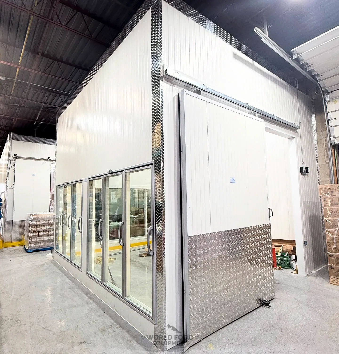Commercial walk-in cooler with a row of black-framed glass-door reach-in display merchandisers built into the front face, white insulated panel construction with brushed aluminum diamond-plate corner trim and kick plates, a solid swing access door alongside, glass-door reach-in cooler installation by World Food Equipment in Ontario