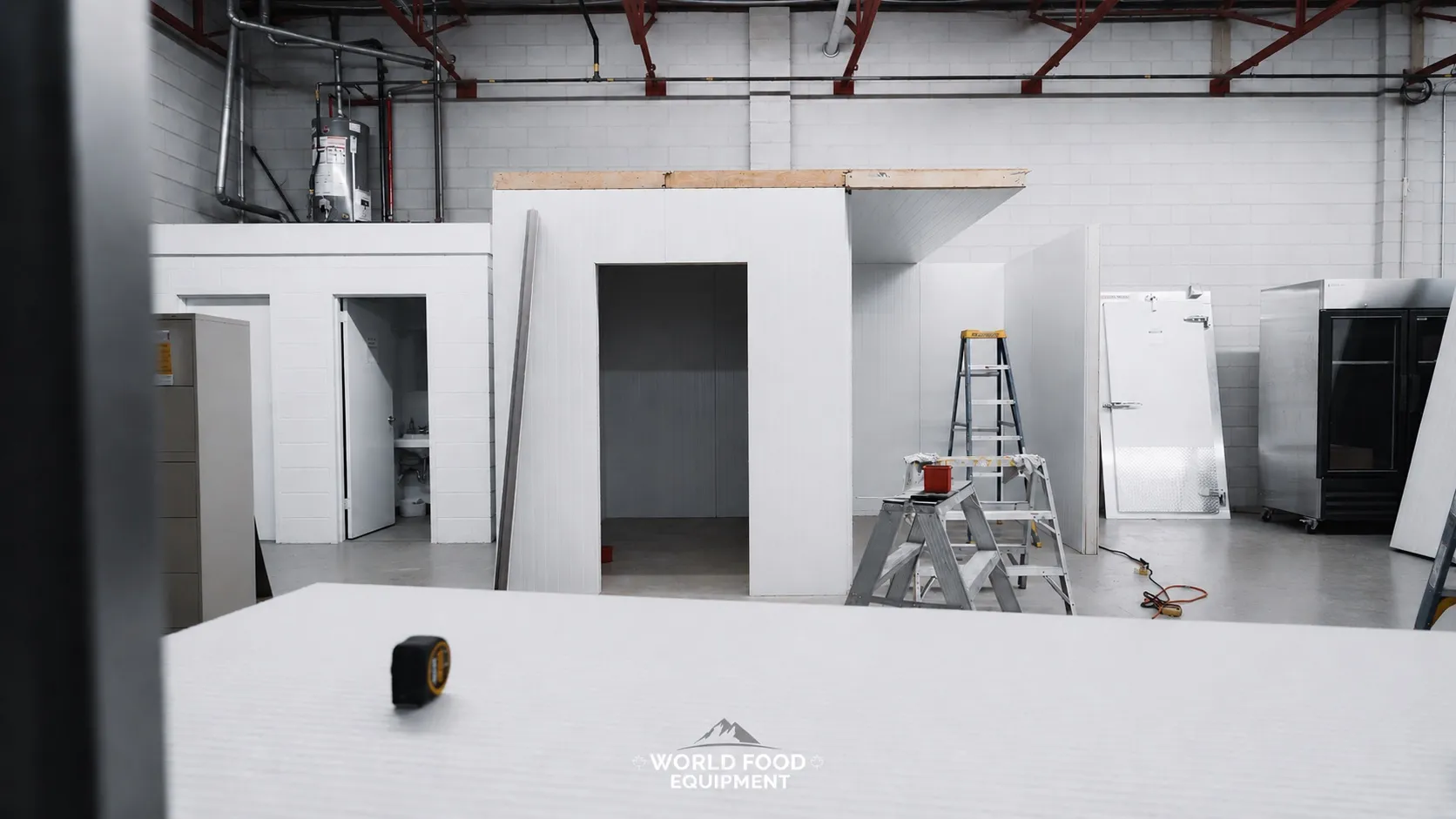 Wide interior view of a commercial warehouse with multiple finished stand-alone walk-in cooler envelopes installed on the left and center, a wide doorway opening into a finished cold storage room at the center, a tall white insulated reach-in unit and a black commercial reach-in cooler visible on the right, yellow A-frame step ladder and scissor lift staged in the room, foreground panel with a tape measure on top, concrete-block warehouse walls and exposed overhead piping, polished concrete floor, Ontario commercial walk-in and reach-in refrigeration by World Food Equipment