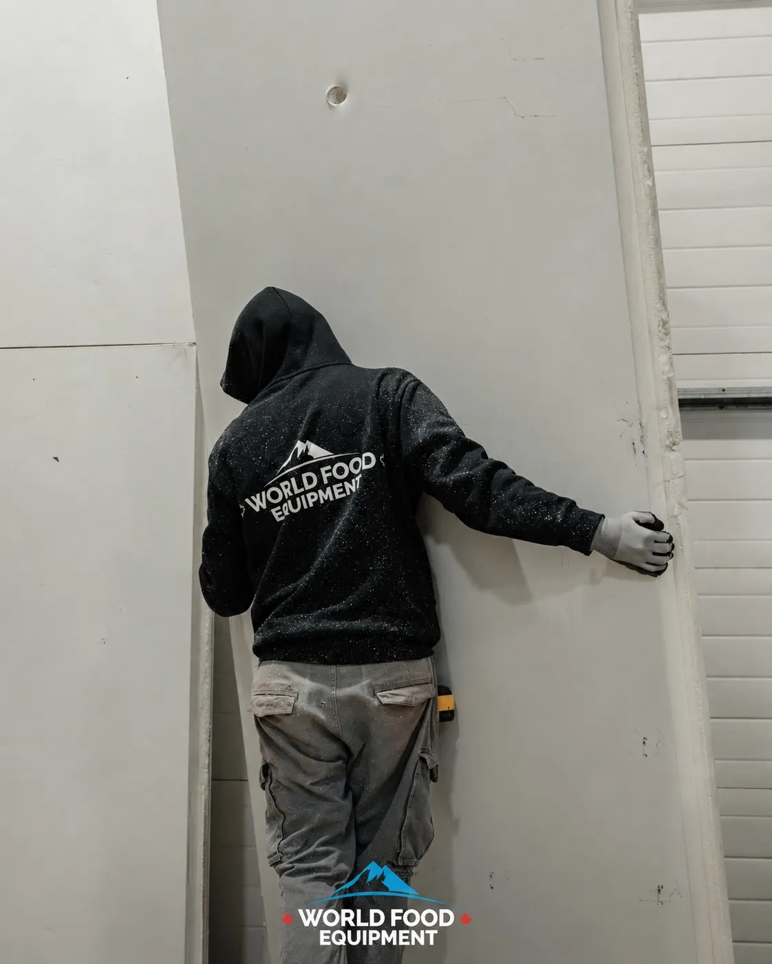 World Food Equipment installer in a branded black WFE hoodie viewed from behind, mountain logo and wordmark centered on the back, gloved hand pressed against the edge of a tall white insulated cold-storage panel during a doorway-frame install, tape measure clipped at the hip, daylight through a corrugated overhead door on the right, polished concrete floor, Ontario commercial walk-in installation by World Food Equipment