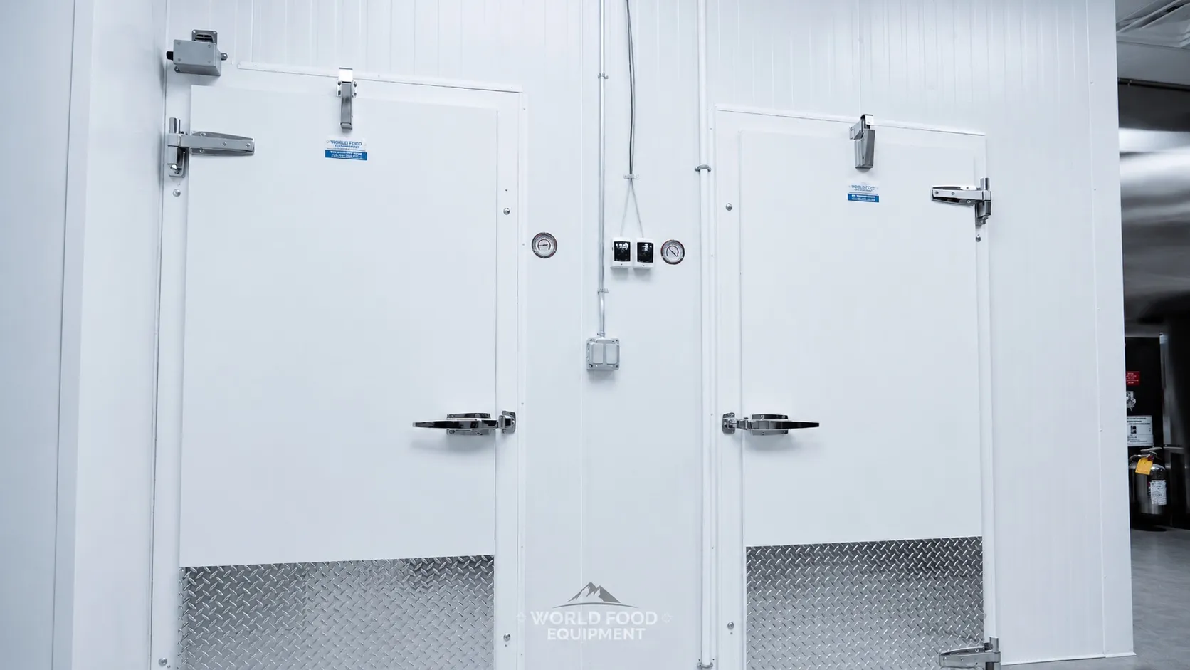 Close-up of two finished commercial walk-in cooler doors with white insulated panel construction, diamond-plate kickplates at the base, dual latch handles on each door, twin NSF stainless dial thermometers mounted at the top of each door, two digital temperature controllers between the doors, Ontario commercial walk-in cooler by World Food Equipment