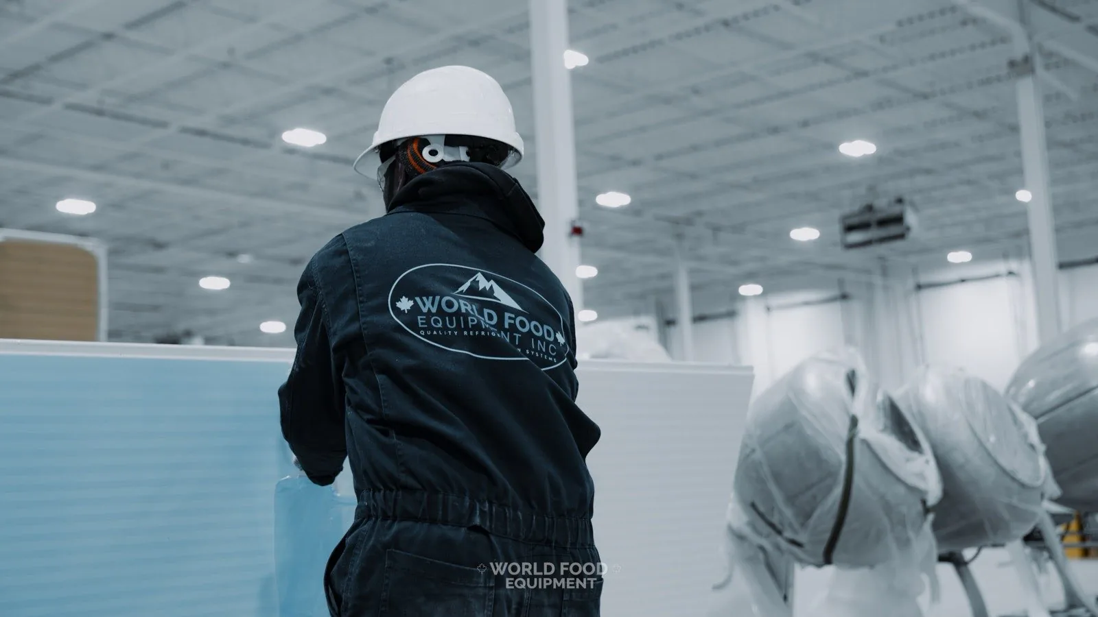 WFE installer in branded coveralls carrying an insulated walk-in freezer panel with wrapped refrigeration equipment staged behind him at an Ontario build